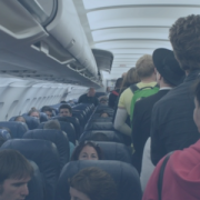 image of people walking in the aisle of a plane and finding their seats. Many people are already sitting. The image has a transparent blue filter over it
