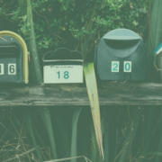 Image of a row of 6 different mailboxes. Behind the mailboxes are plants and foliage.